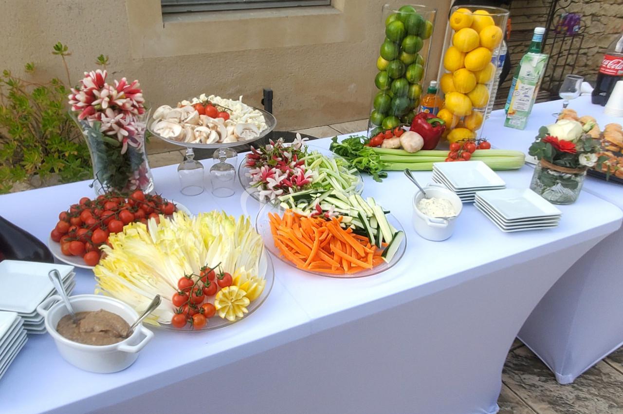 Buffet en plein air avec légumes frais, fruits, sauces et pains disposés sur une table blanche.