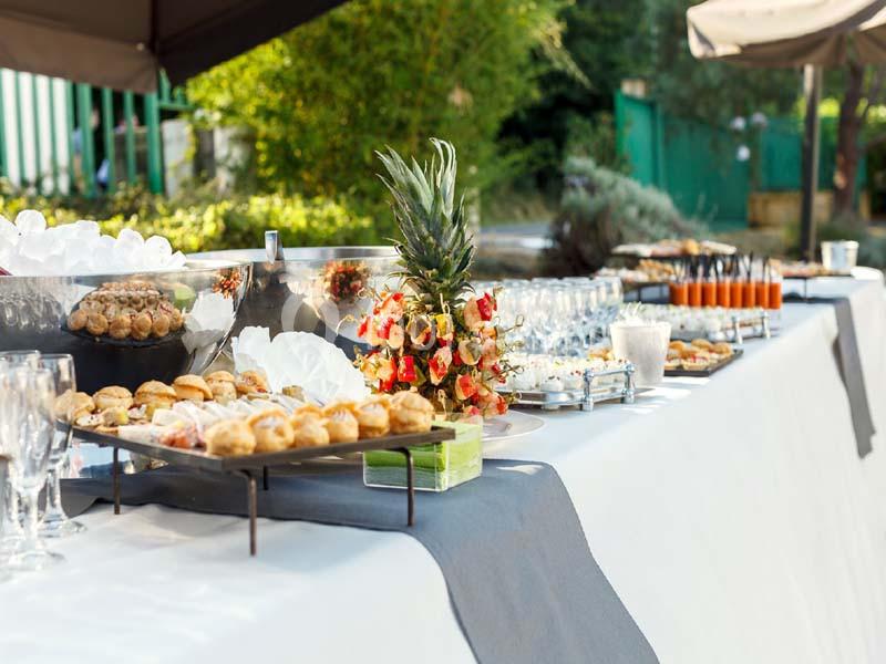 Buffet en extérieur avec divers plats, verrines, brochettes de fruits et boissons disposés sur une table blanche.