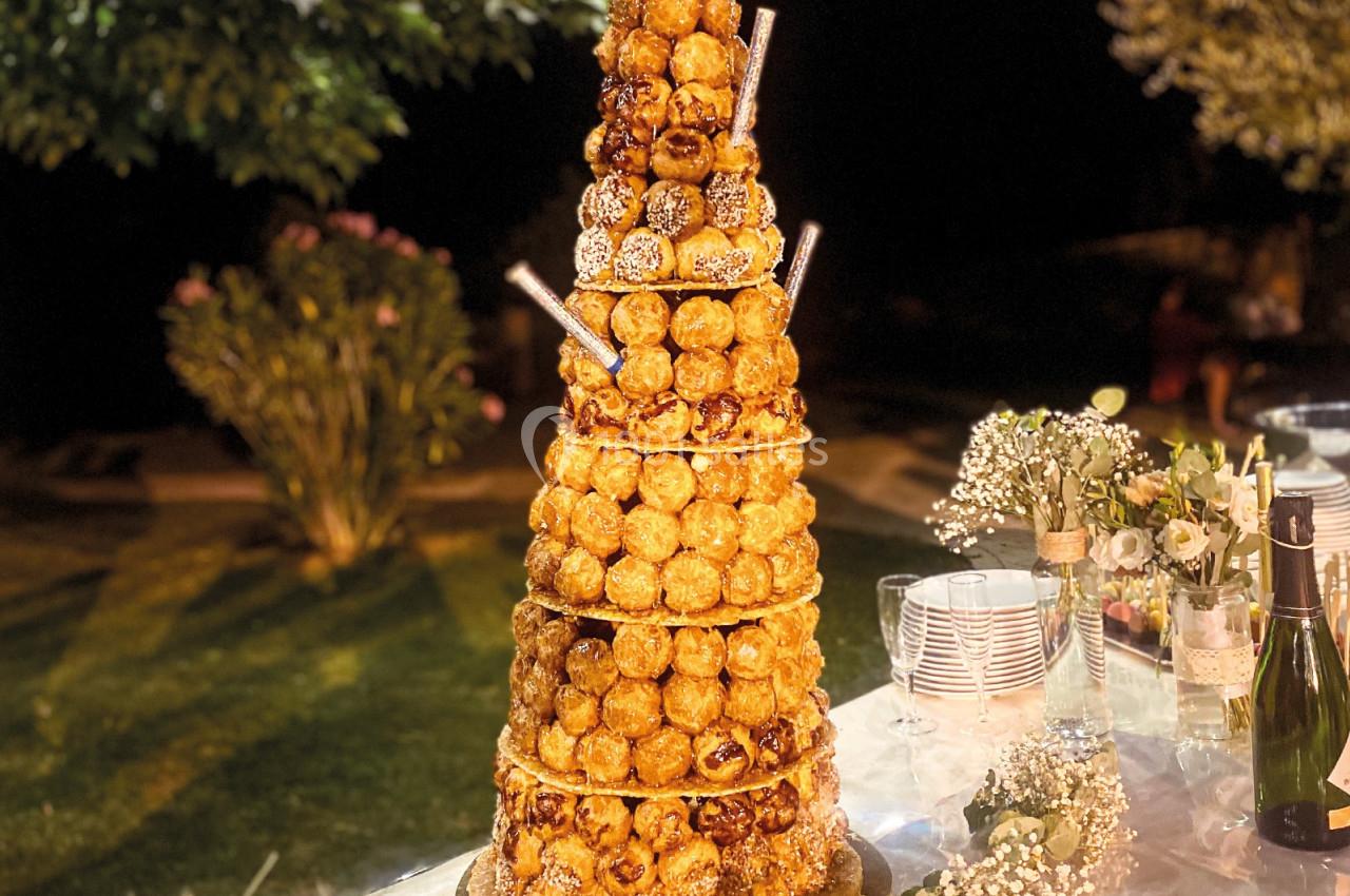 Tour de choux en croquembouche décorée, posée sur une table extérieure éclairée, entourée de fleurs et de vaisselle.