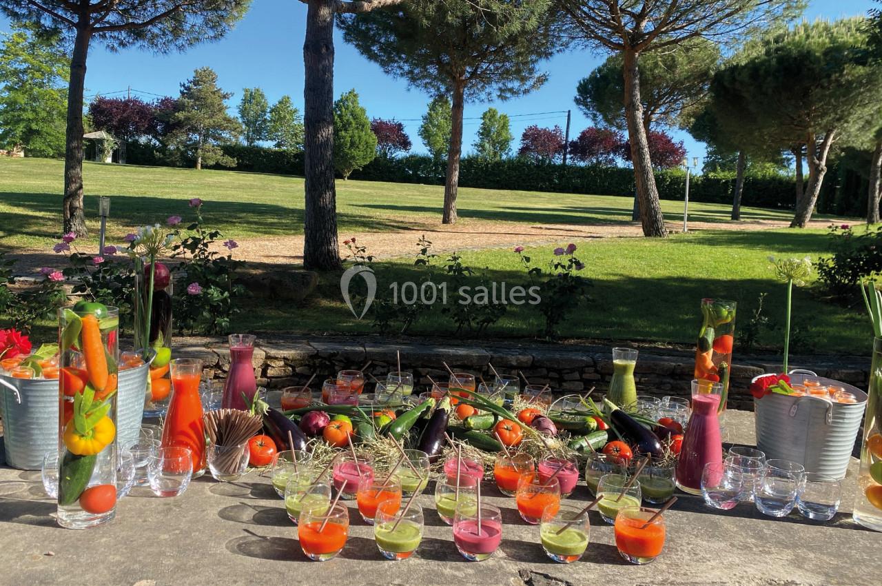Buffet en plein air avec verrines colorées, légumes frais et jus, entouré de verdure et d'arbres sous un ciel bleu.