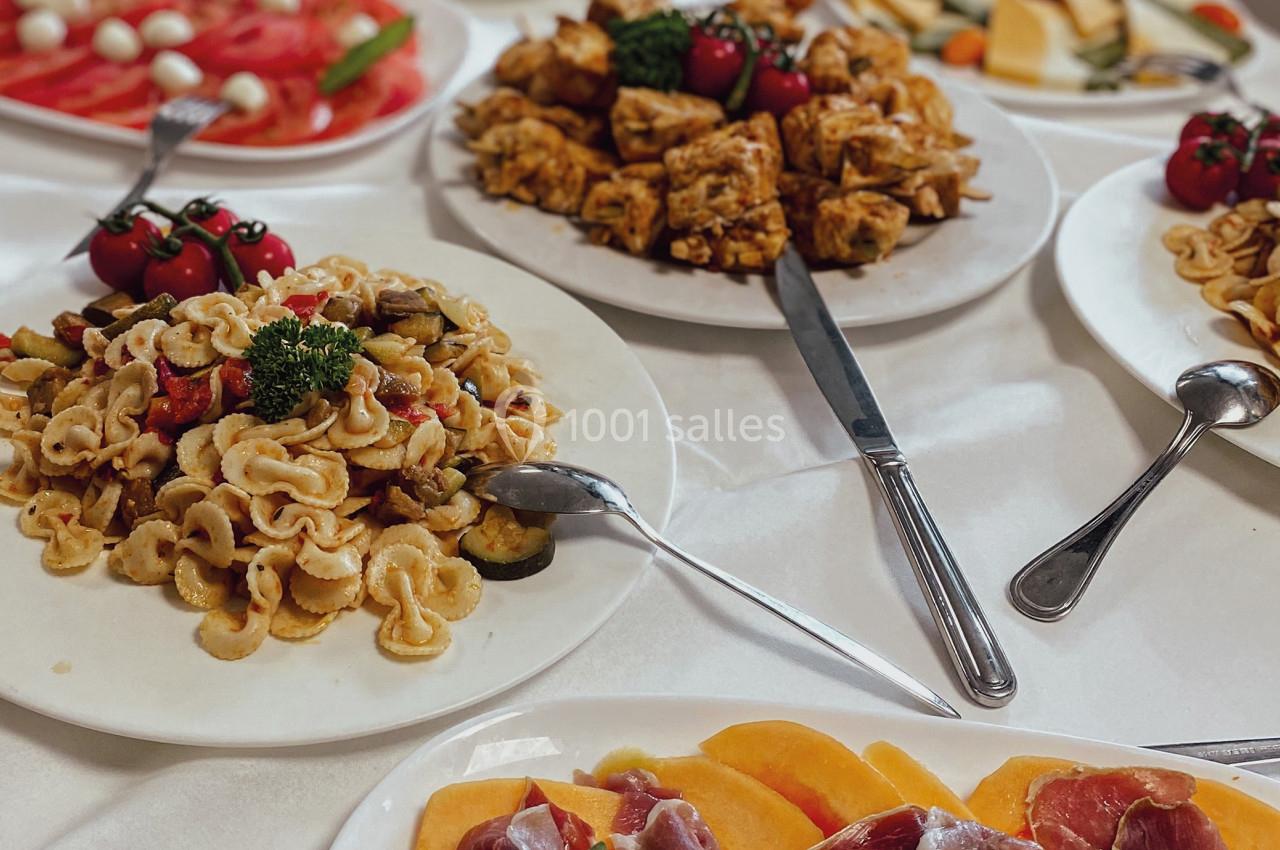 Assortiment de plats variés comprenant des pâtes, brochettes, charcuterie et légumes, présentés sur une table blanche.