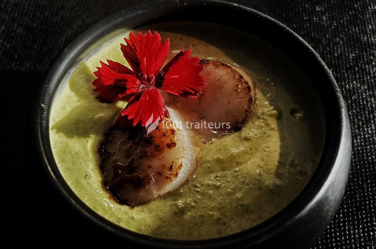 Velouté vert garni de deux noix de Saint-Jacques poêlées et décoré d'une fleur rouge, présenté dans un bol noir.