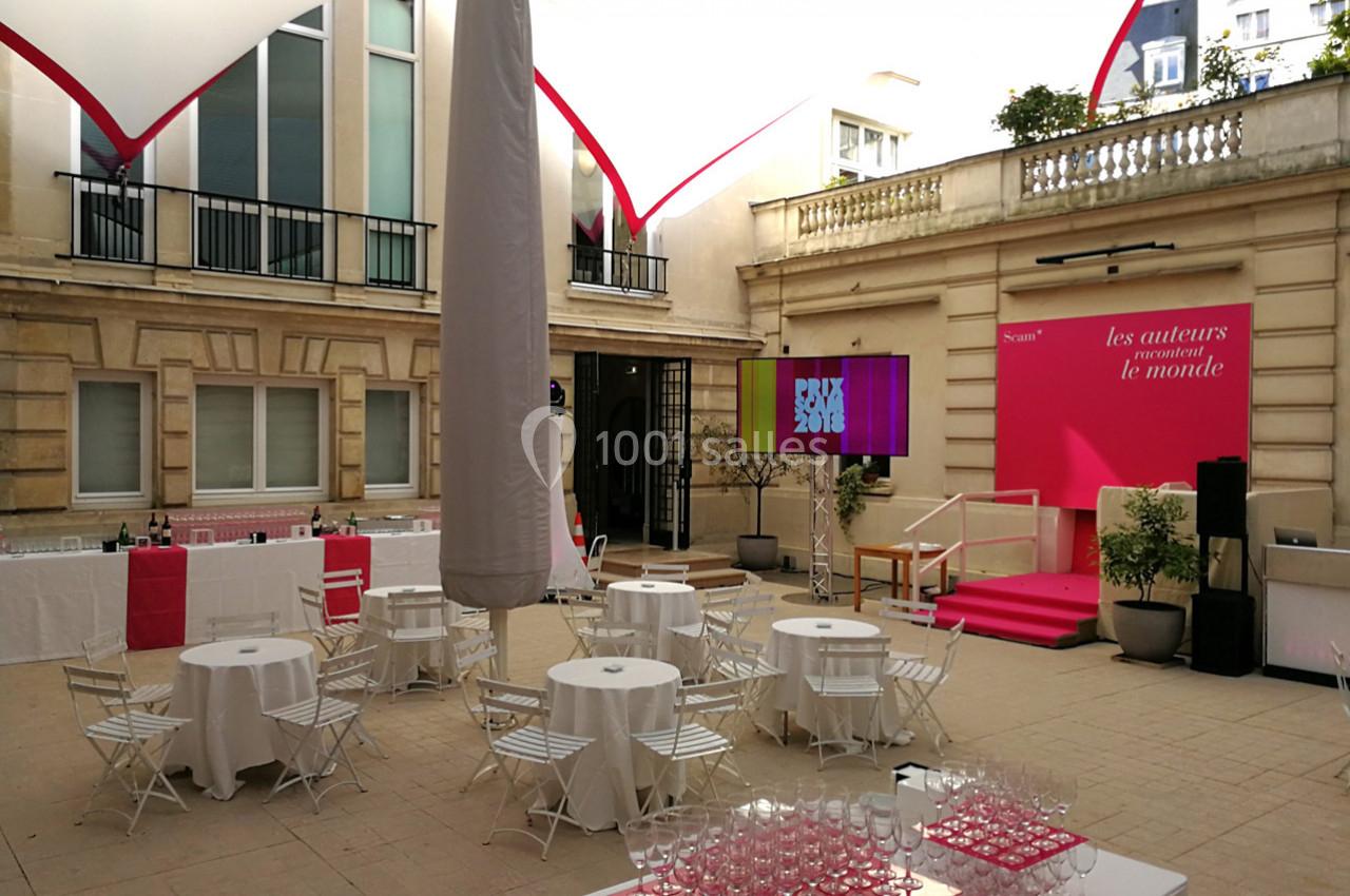 Cour intérieure aménagée pour un événement avec tables rondes, chaises blanches et estrade rose en arrière-plan.