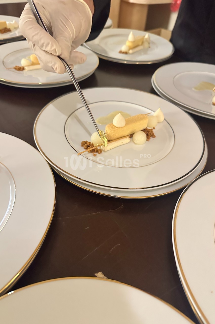 Un chef décore un dessert raffiné avec une pince, sur une assiette blanche bordée d'or.
