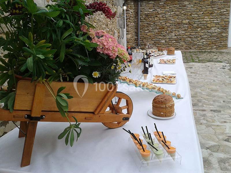 Table dressée en extérieur avec des amuse-bouches, une charrette fleurie en bois et des boissons sur une nappe blanche.
