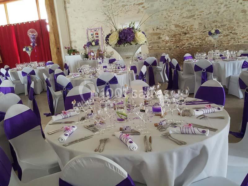 Salle décorée pour un mariage avec des tables rondes dressées, nappes blanches et accents violets.