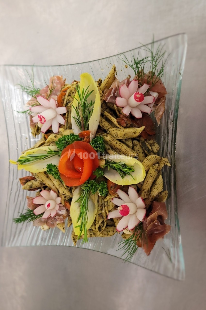 Assiette de pâtes aux herbes, décorée de légumes sculptés, feuilles d'endive, jambon cru et herbes fraîches.
