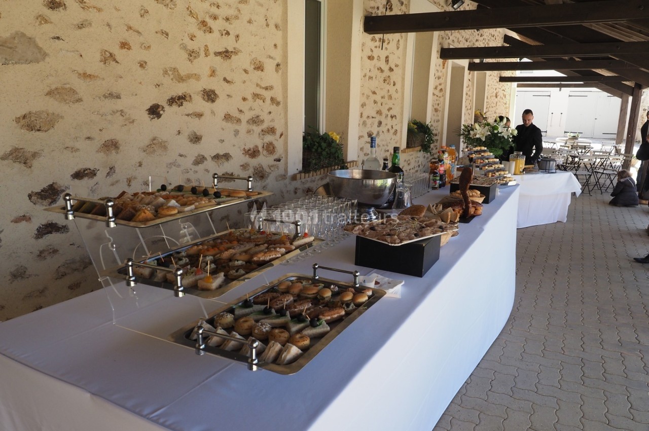 Buffet en extérieur avec viennoiseries, desserts, boissons et vaisselle, disposé sur une table blanche sous un auvent.