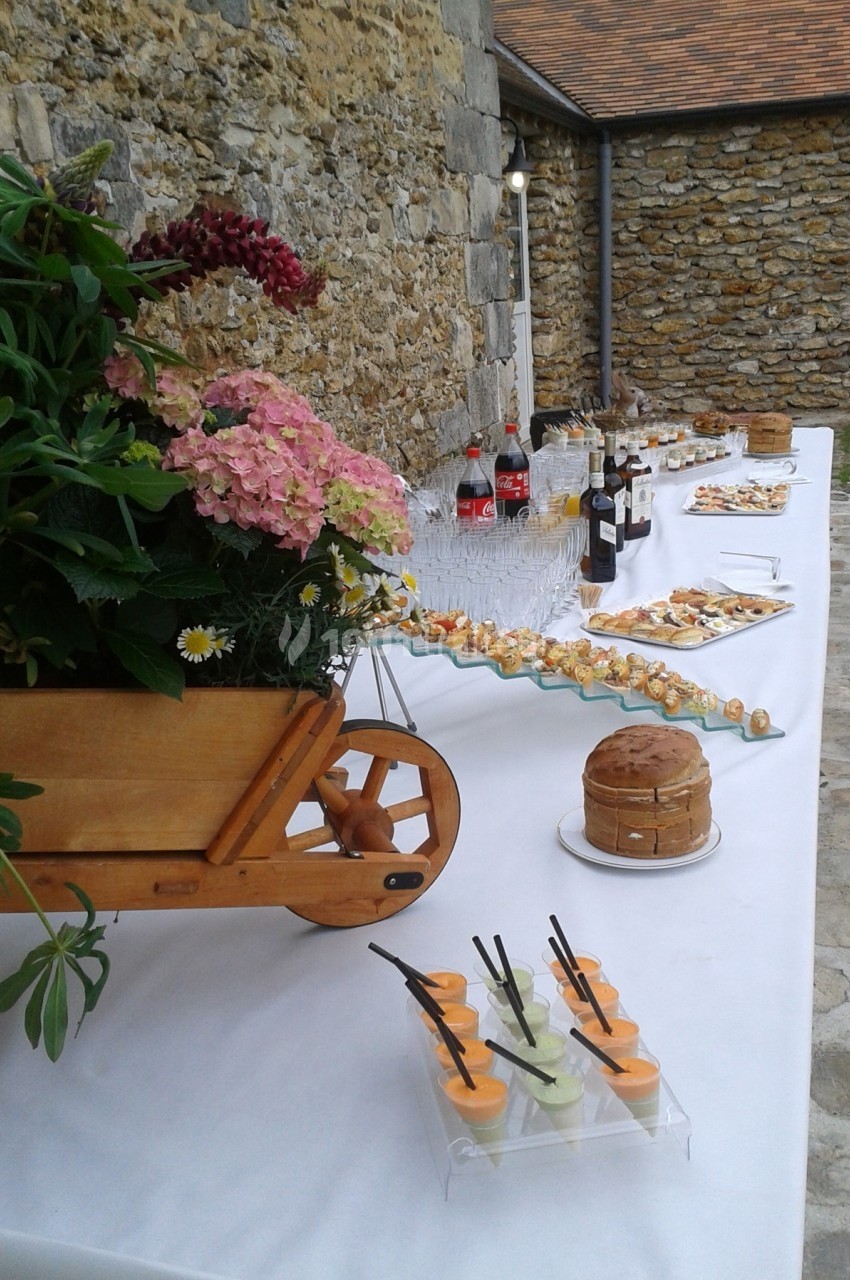 Table de buffet en extérieur avec boissons, verrines, amuse-bouches, gâteau et décoration florale dans une brouette en bois.