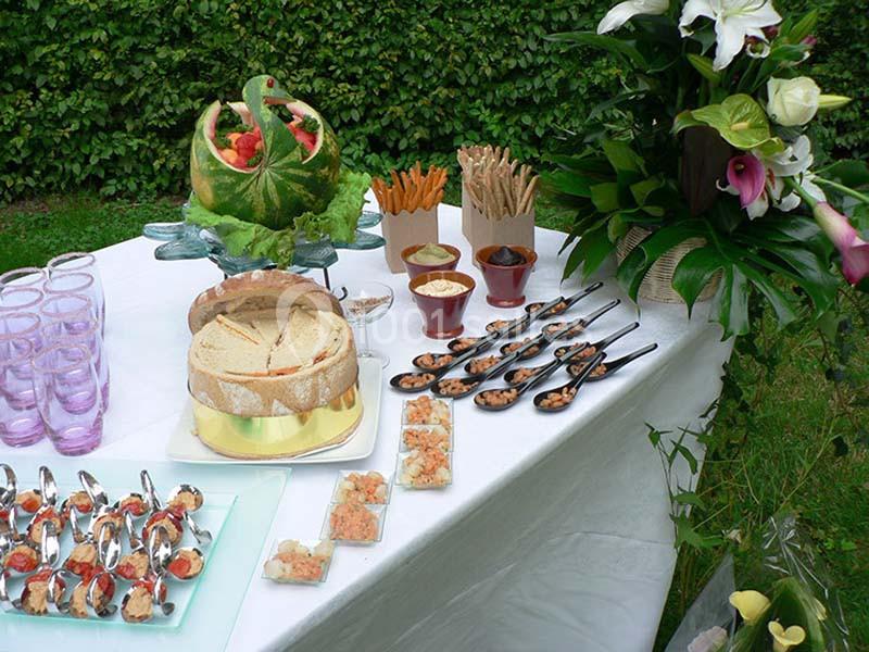 Table de buffet en extérieur avec pain, verrines, cuillères apéritives, fruits sculptés et fleurs décoratives.