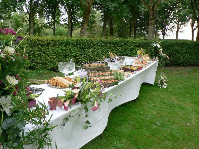 Table de buffet dressée en extérieur, garnie de plats variés, fleurs et verrerie, entourée de verdure et d'arbres.