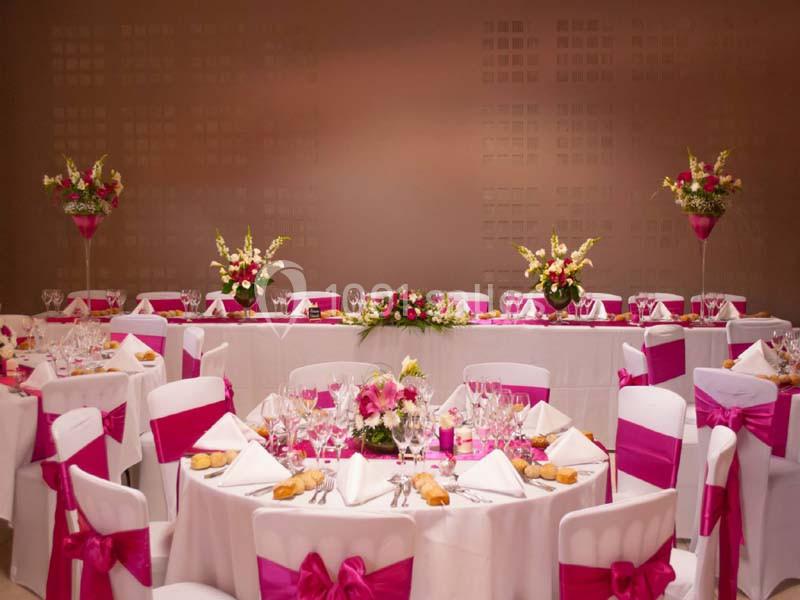 Salle de réception décorée avec des nappes blanches, des nœuds roses sur les chaises et des compositions florales colorées.