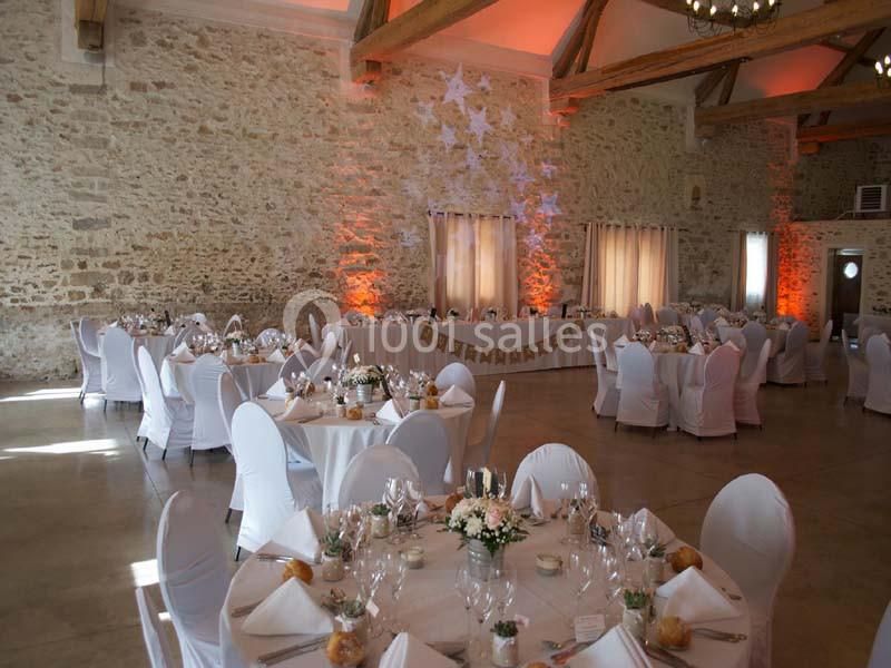 Salle de réception avec murs en pierre, tables rondes décorées et chaises blanches, éclairage chaleureux.