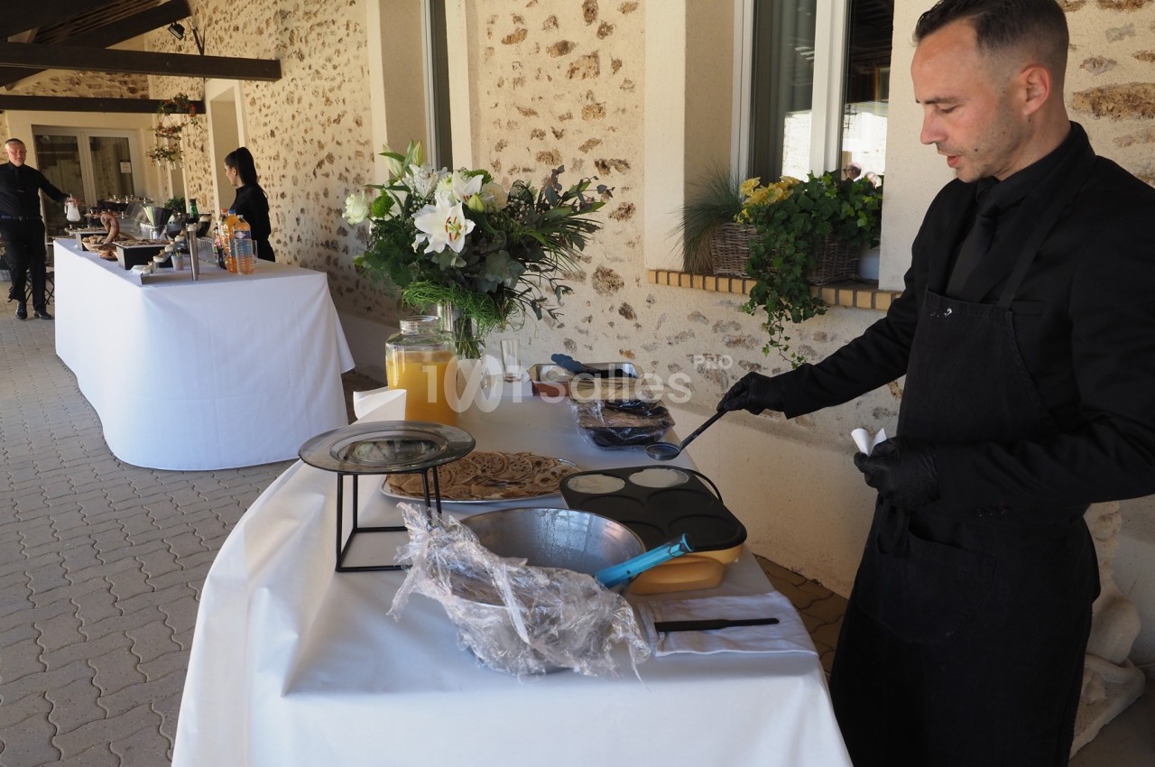 Un cuisinier prépare des crêpes sur une plaque chauffante lors d'un buffet en extérieur près d'un bâtiment en pierre.