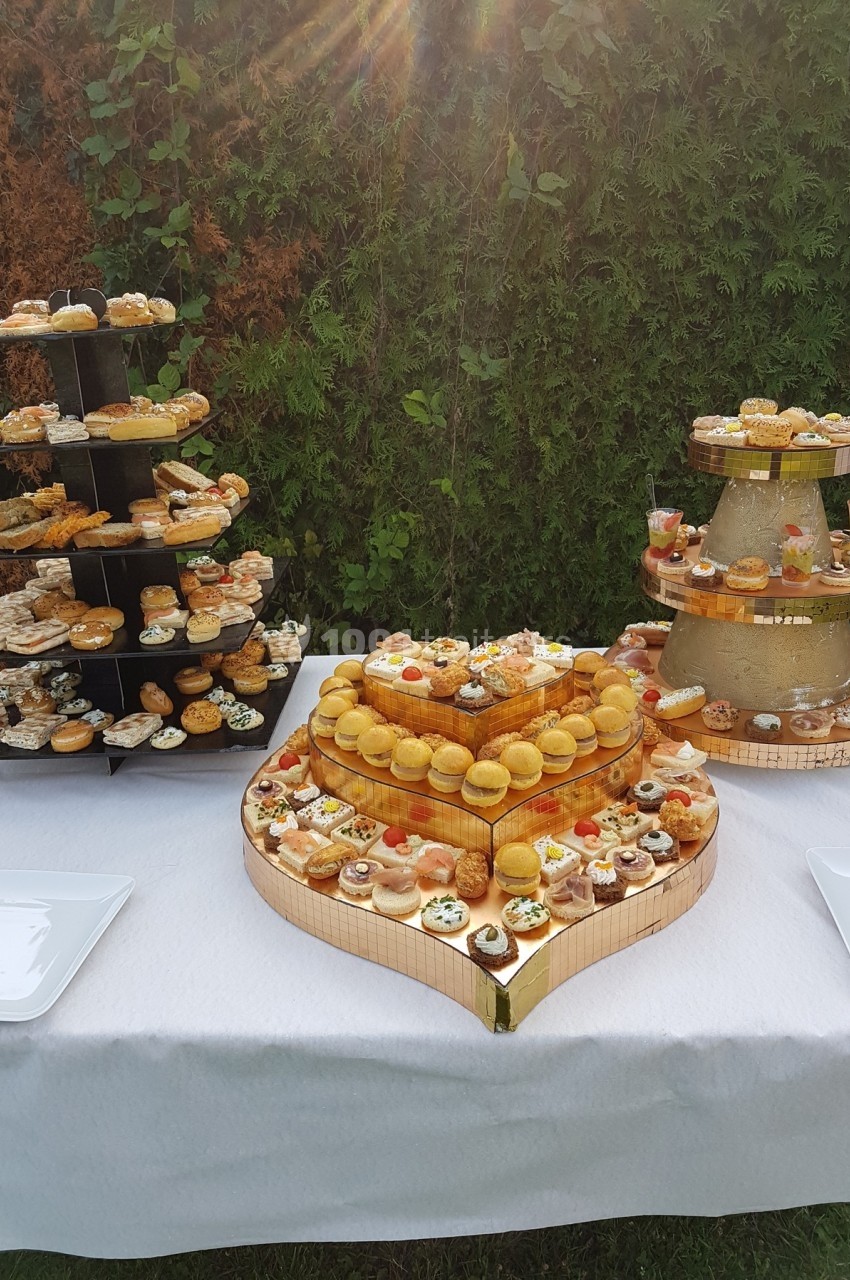 Buffet de pâtisseries et amuse-bouches disposés sur des présentoirs en extérieur devant une haie verte.