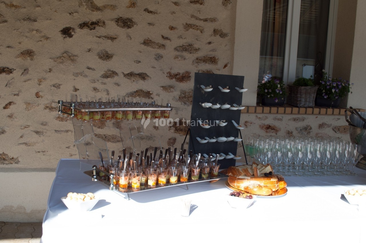 Buffet en extérieur avec verrines, cuillères apéritives, pain, viennoiseries et verres alignés sur une table blanche.