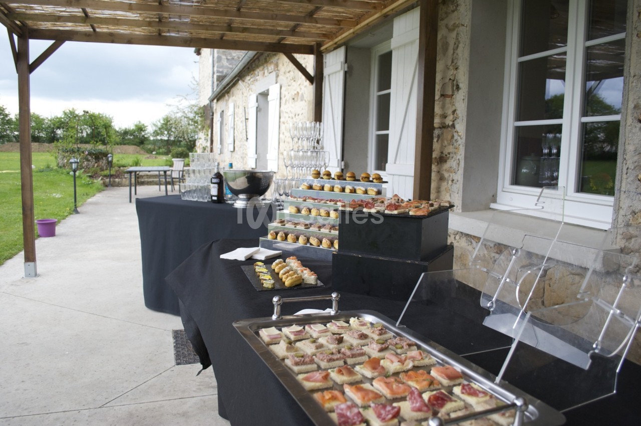 Buffet en extérieur sous une pergola, avec plateaux de canapés, verrines et gâteaux disposés sur des nappes noires.