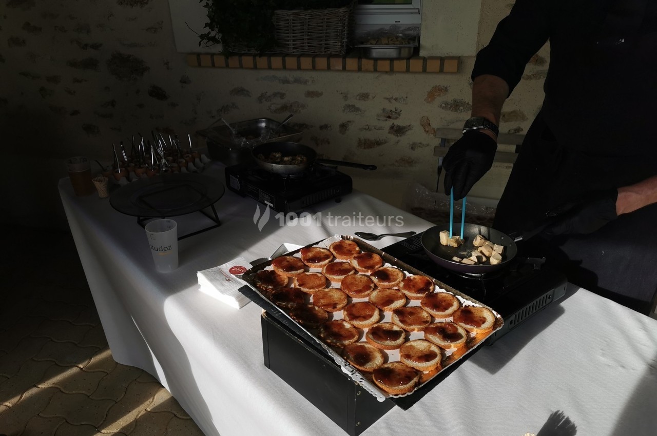 Un cuisinier prépare des aliments dans une poêle à côté d'un plateau de mini-tartes sur une table blanche.