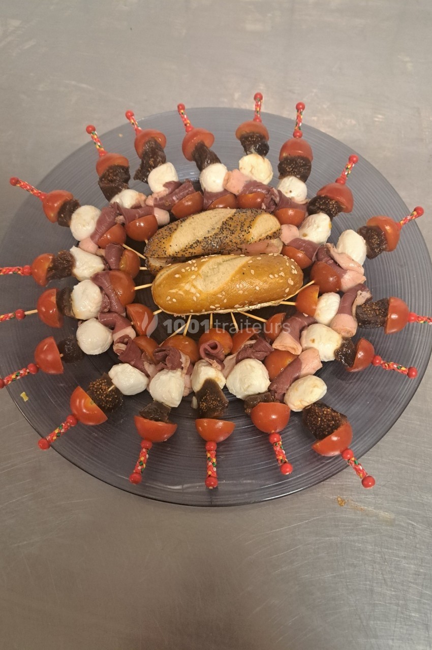 Assiette circulaire garnie de brochettes apéritives avec tomates, mozzarella, charcuterie et pains au centre.