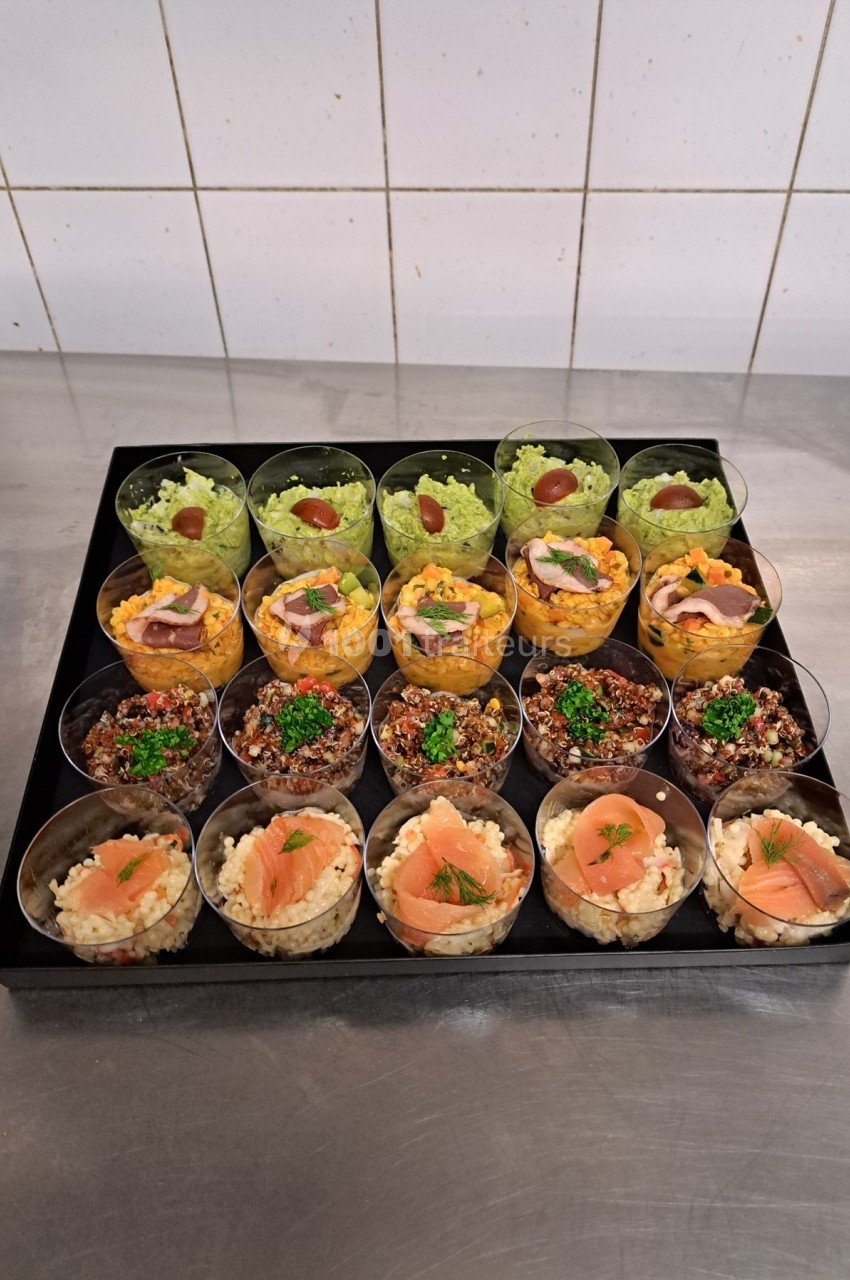 Plateau de verrines variées contenant des préparations à base de légumes, quinoa, saumon et purée, sur un plan de travail.