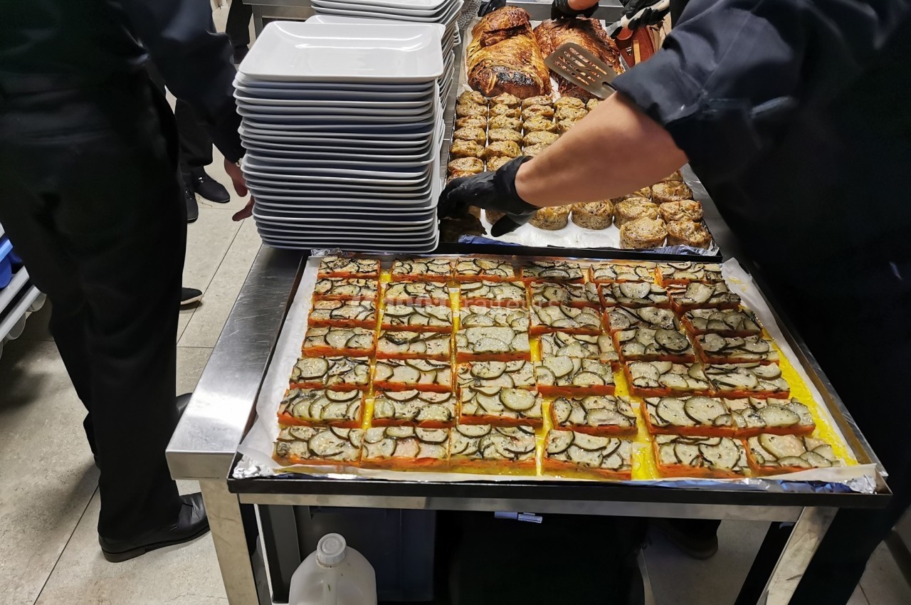 Des cuisiniers préparent des plats en cuisine, avec des assiettes empilées et une grande plaque de quiches aux légumes.