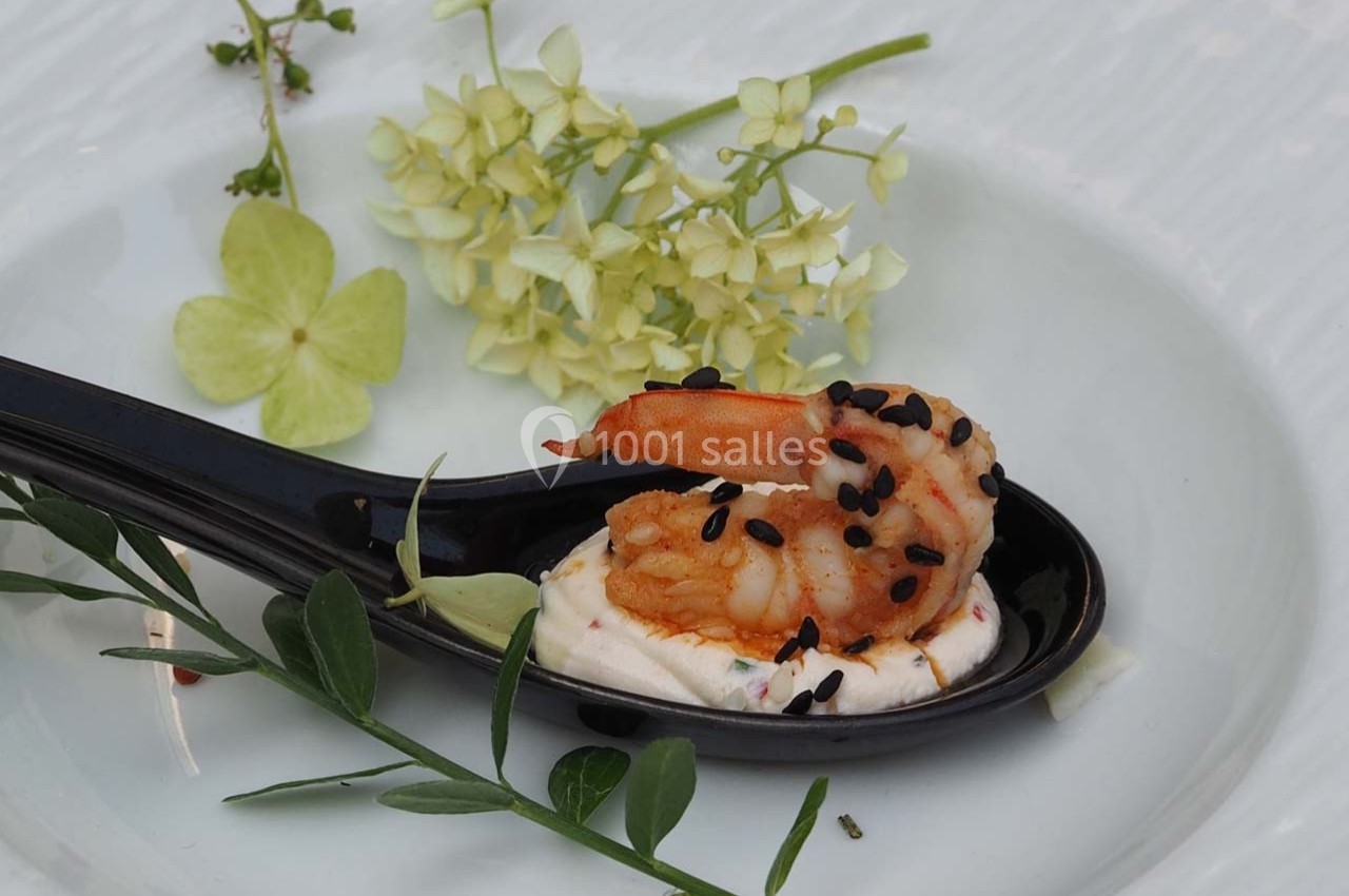 Crevette grillée sur une cuillère noire avec une base de crème, décorée de graines de sésame et de fleurs.