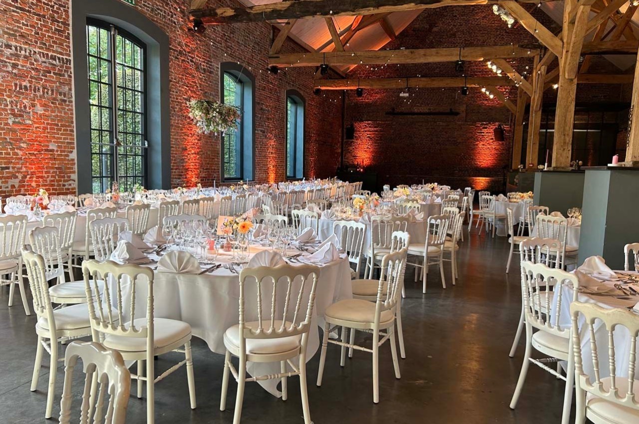 Salle de réception avec murs en briques, tables rondes dressées et chaises blanches, éclairage chaleureux.