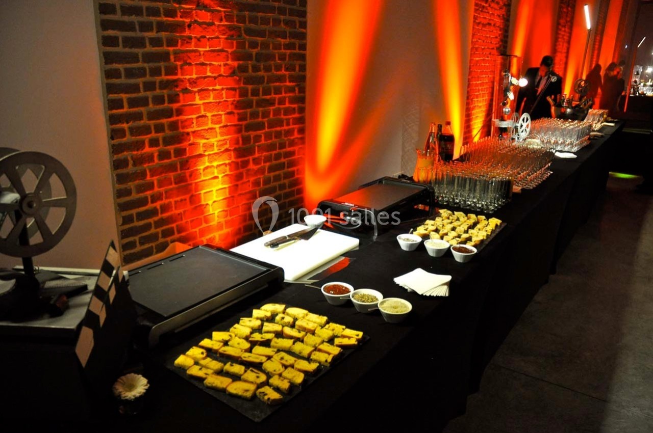 Table de buffet avec planchas, verrines, sauces et amuse-bouches, éclairée par des lumières orangées près d'un mur en…