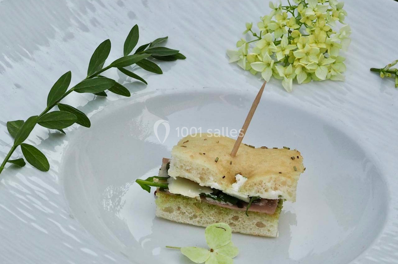 Mini-sandwich sur pain focaccia garni de fromage, jambon et roquette, présenté sur une assiette blanche avec décor floral.