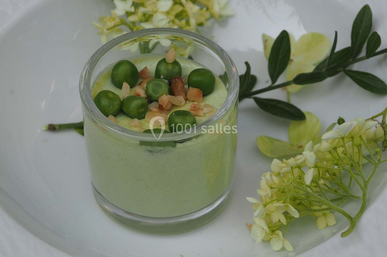 Verrine de mousse verte garnie de petits pois et éclats de noix, entourée de fleurs et de feuillage sur une assiette blanche.