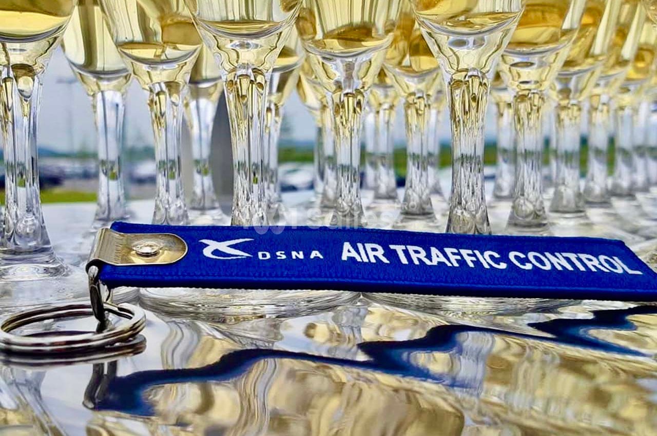 Rangée de flûtes de champagne alignées sur une table avec un porte-clés bleu marqué ’DSNA Air Traffic Control’.