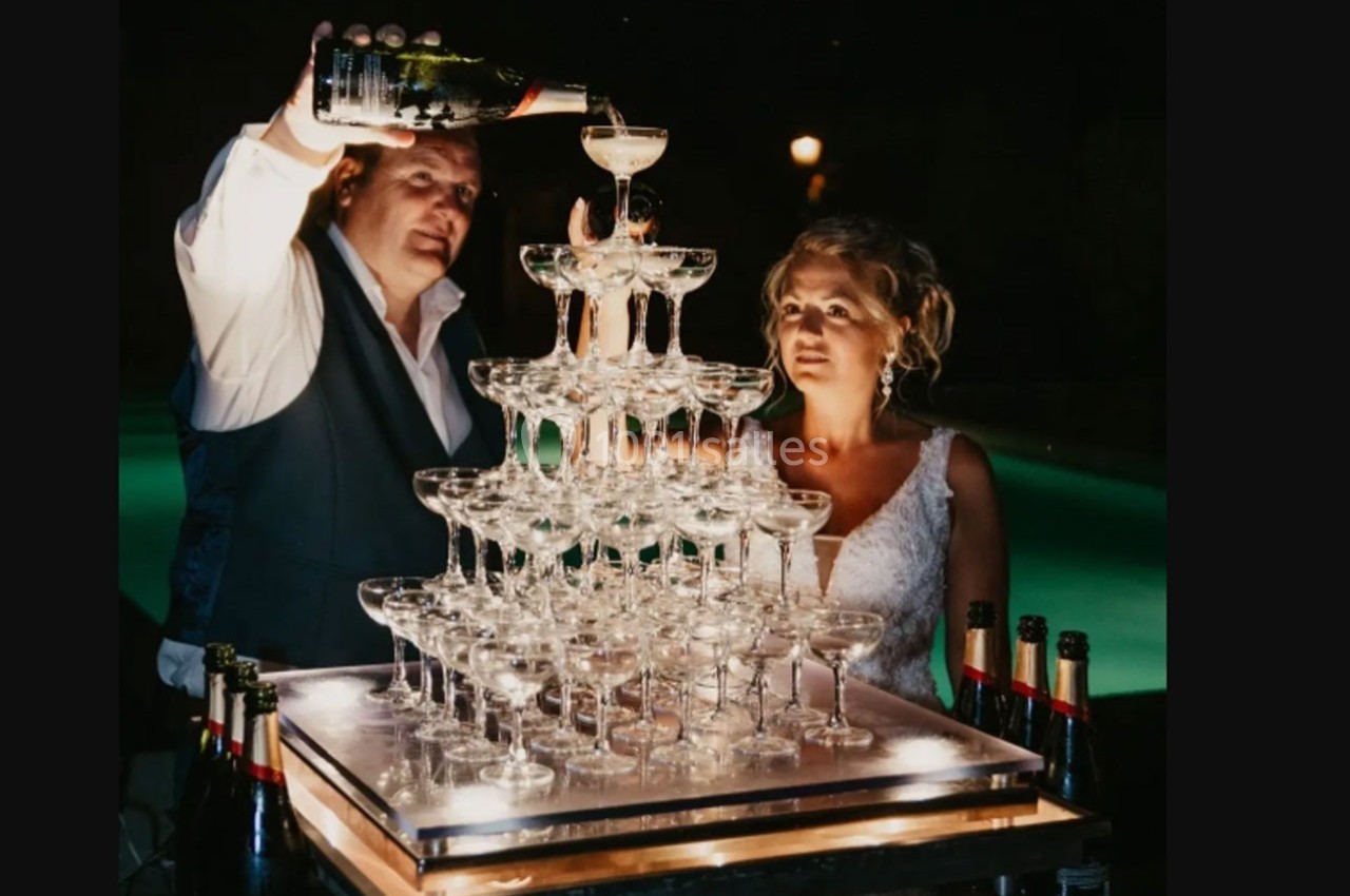 Un homme verse du champagne sur une pyramide de coupes en verre, accompagné d'une femme en robe blanche.