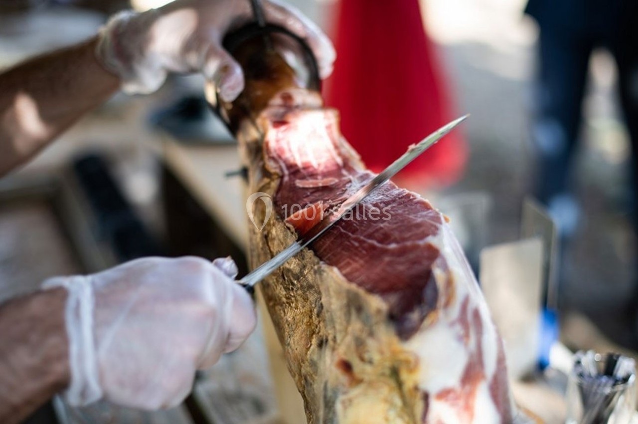 Découpe d'une tranche de jambon cru sur un support, réalisée avec un couteau par une personne portant des gants.