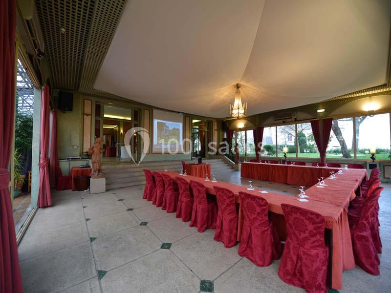 Salle de réunion lumineuse avec tables en U couvertes de nappes rouges, ouverte sur un jardin verdoyant.