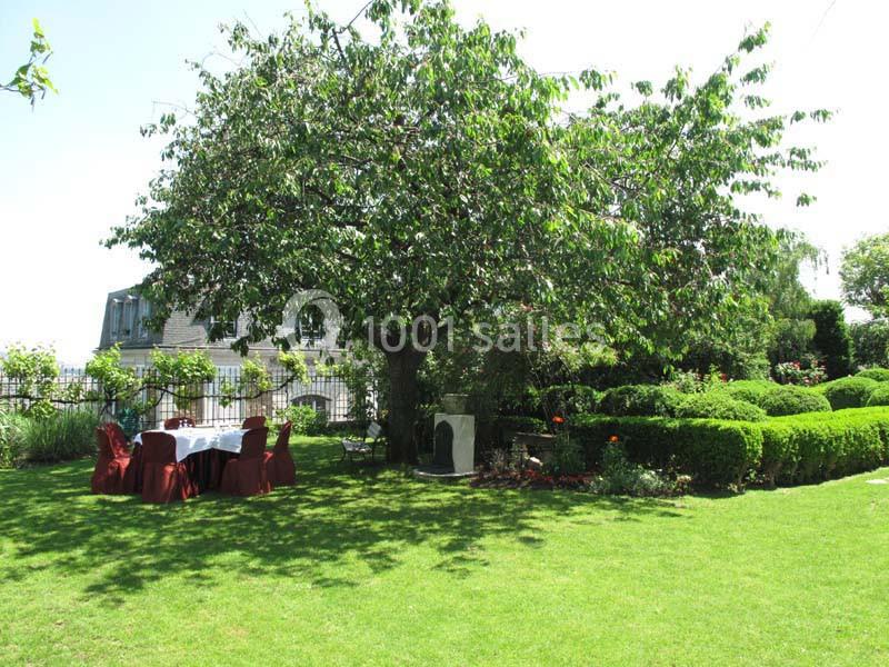 Un jardin verdoyant avec un grand arbre ombrageant une table dressée et des chaises rouges.