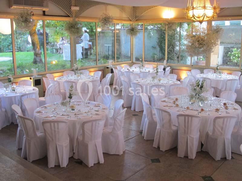Salle de réception lumineuse avec des tables rondes dressées, nappes blanches et vue sur un jardin extérieur.