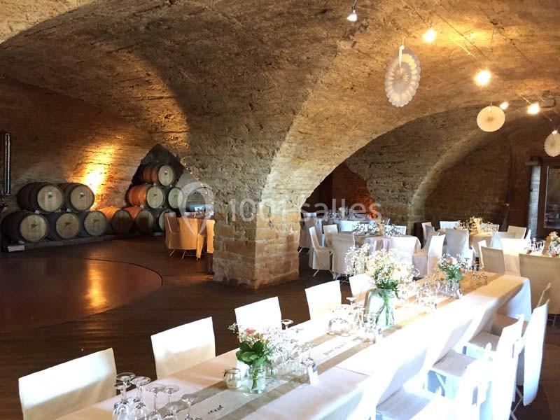 Salle voûtée en pierre aménagée pour un événement, avec tables dressées et tonneaux en arrière-plan.
