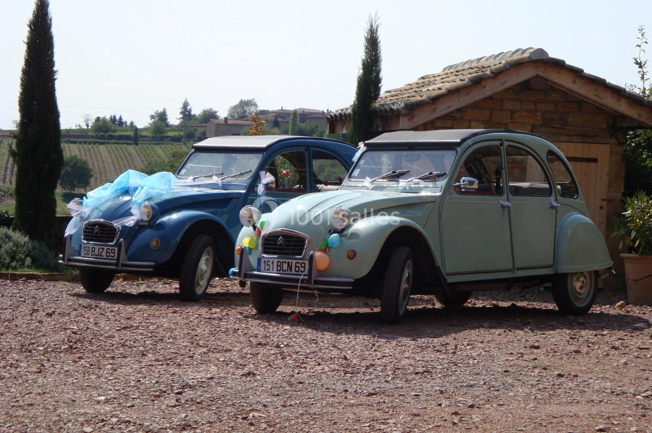 Deux voitures Citroën 2CV, l'une bleue et l'autre verte, stationnées sur un terrain en gravier devant un bâtiment en pierre.