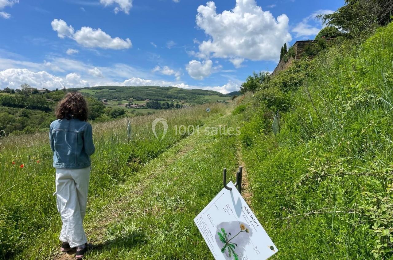 Une femme marche sur un sentier bordé de verdure et de panneaux explicatifs dans un paysage vallonné.