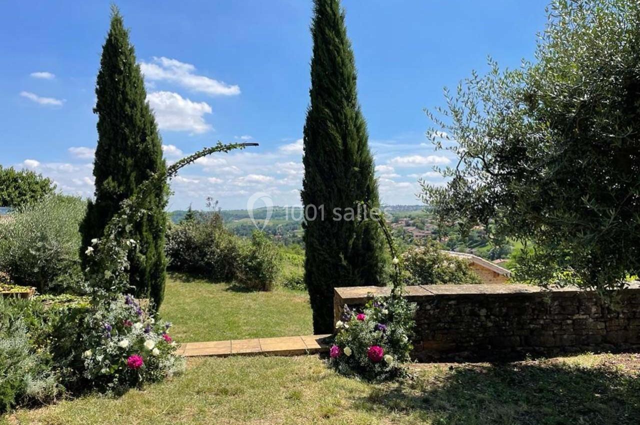 Allée en pierre bordée de cyprès et de fleurs, donnant sur un paysage vallonné sous un ciel bleu.