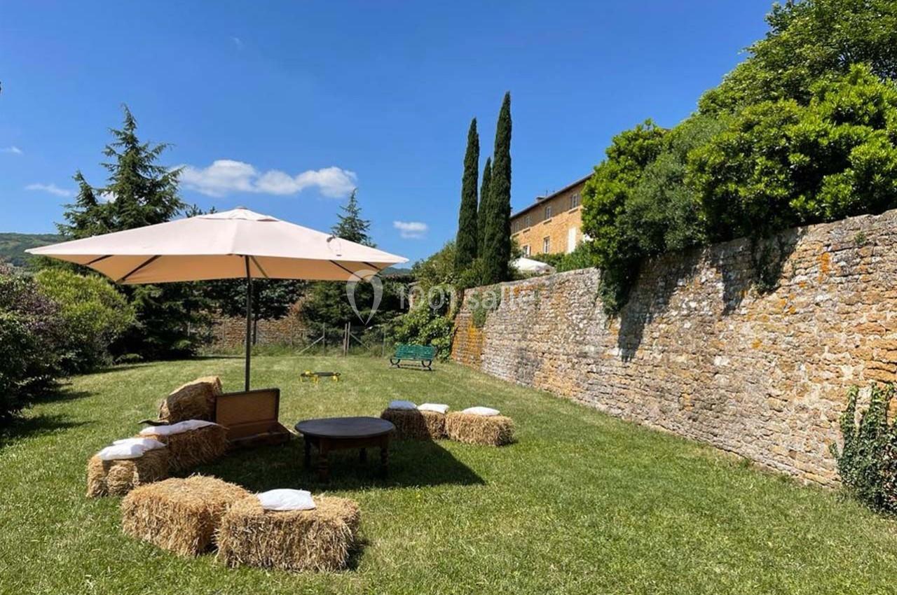 Espace extérieur avec parasol, bottes de paille en sièges, table basse, pelouse et mur en pierre sous un ciel bleu.