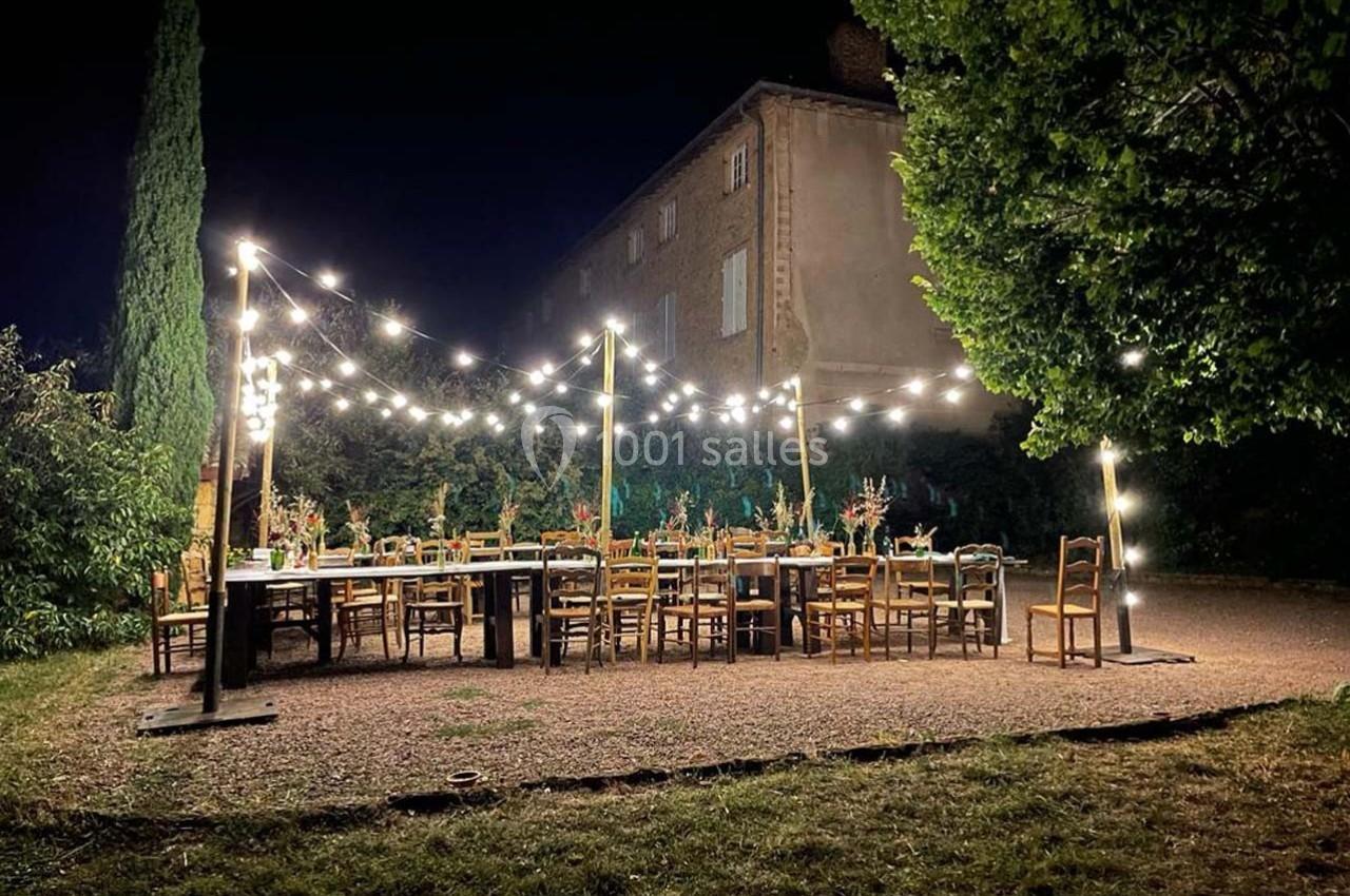 Grande table dressée en extérieur sous des guirlandes lumineuses, entourée de chaises, devant un bâtiment éclairé la nuit.