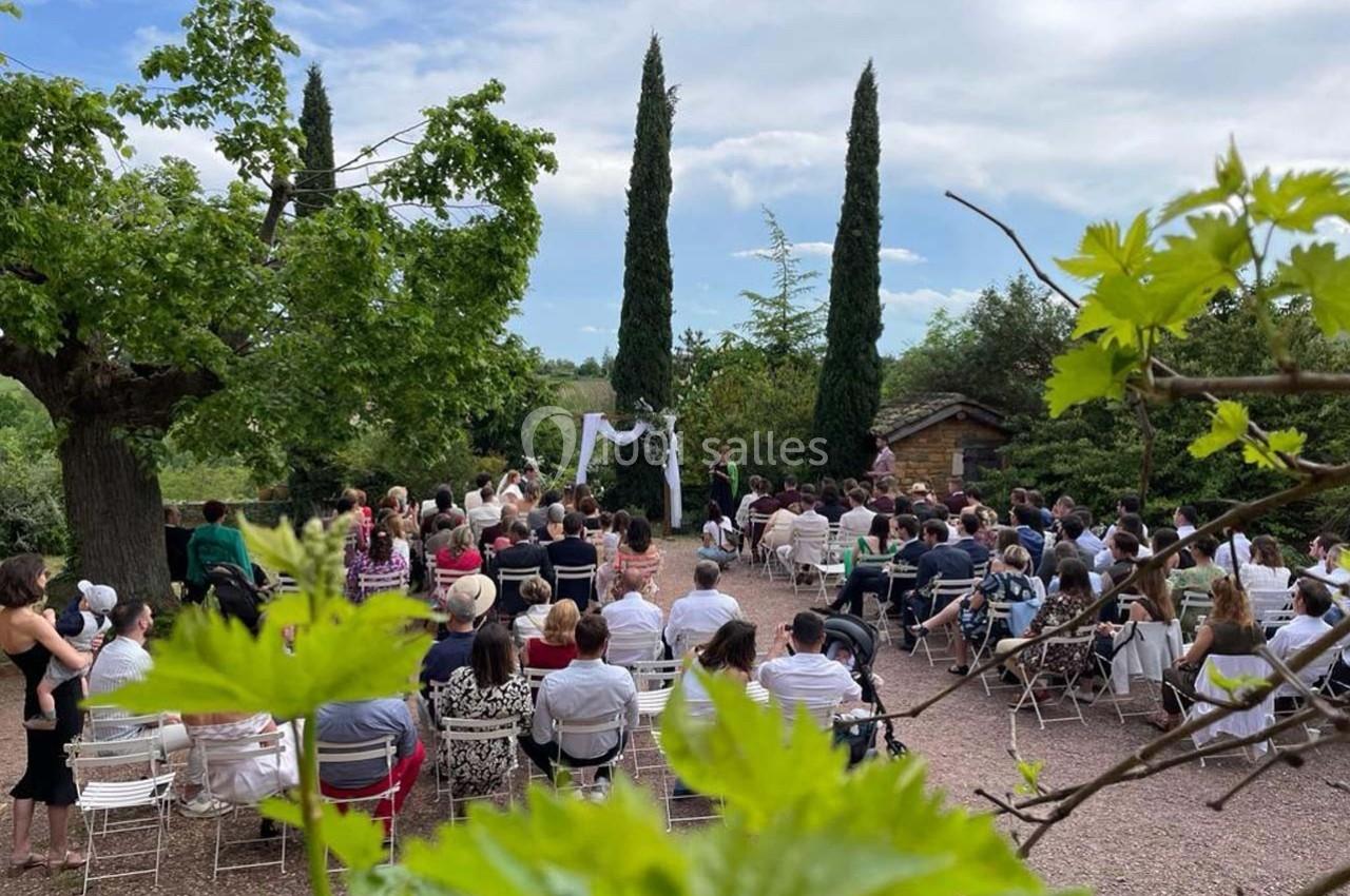 Cérémonie en plein air avec des invités assis, entourée de verdure et de cyprès sous un ciel partiellement nuageux.