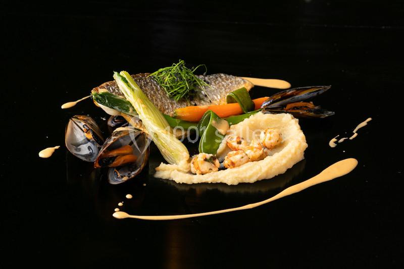 Assiette gastronomique composée de poisson, légumes, moules et purée, présentée sur un fond noir.