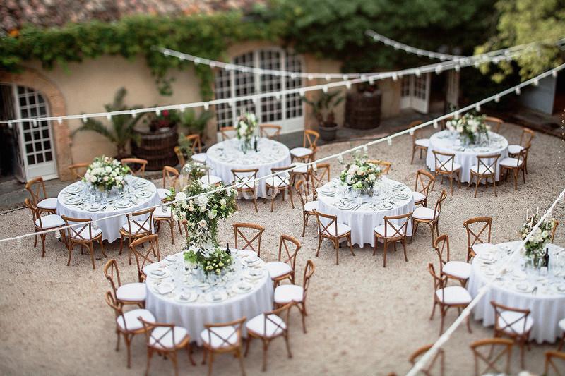 Tables rondes décorées pour un événement en extérieur, entourées de chaises en bois et guirlandes suspendues.