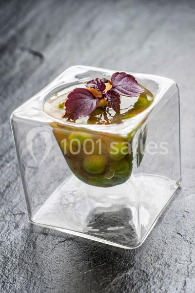 Petit plat gastronomique dans un récipient en verre, contenant des légumes verts et décoré de feuilles pourpres.