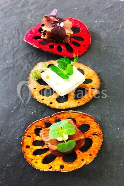 Tranches de lotus colorées garnies de fromage, viande et herbes fraîches, présentées sur une ardoise sombre.