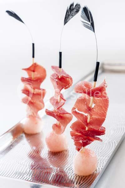 Tranches fines de jambon enroulées sur des brochettes, présentées verticalement sur des supports sphériques.