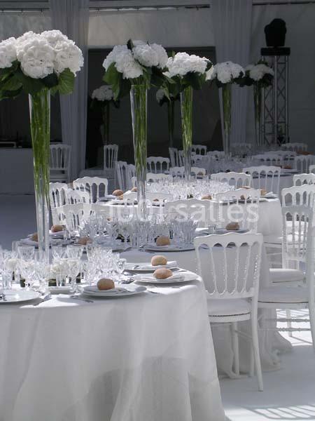 Tables rondes dressées avec nappes blanches, chaises assorties et centres de table composés de fleurs blanches.