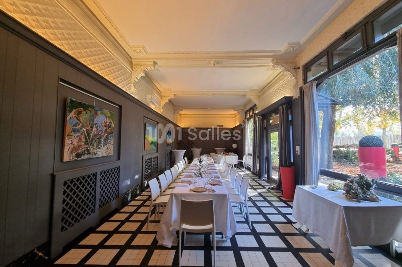 Salle lumineuse avec tables dressées en enfilade, grandes baies vitrées, œuvres d'art accrochées aux murs.